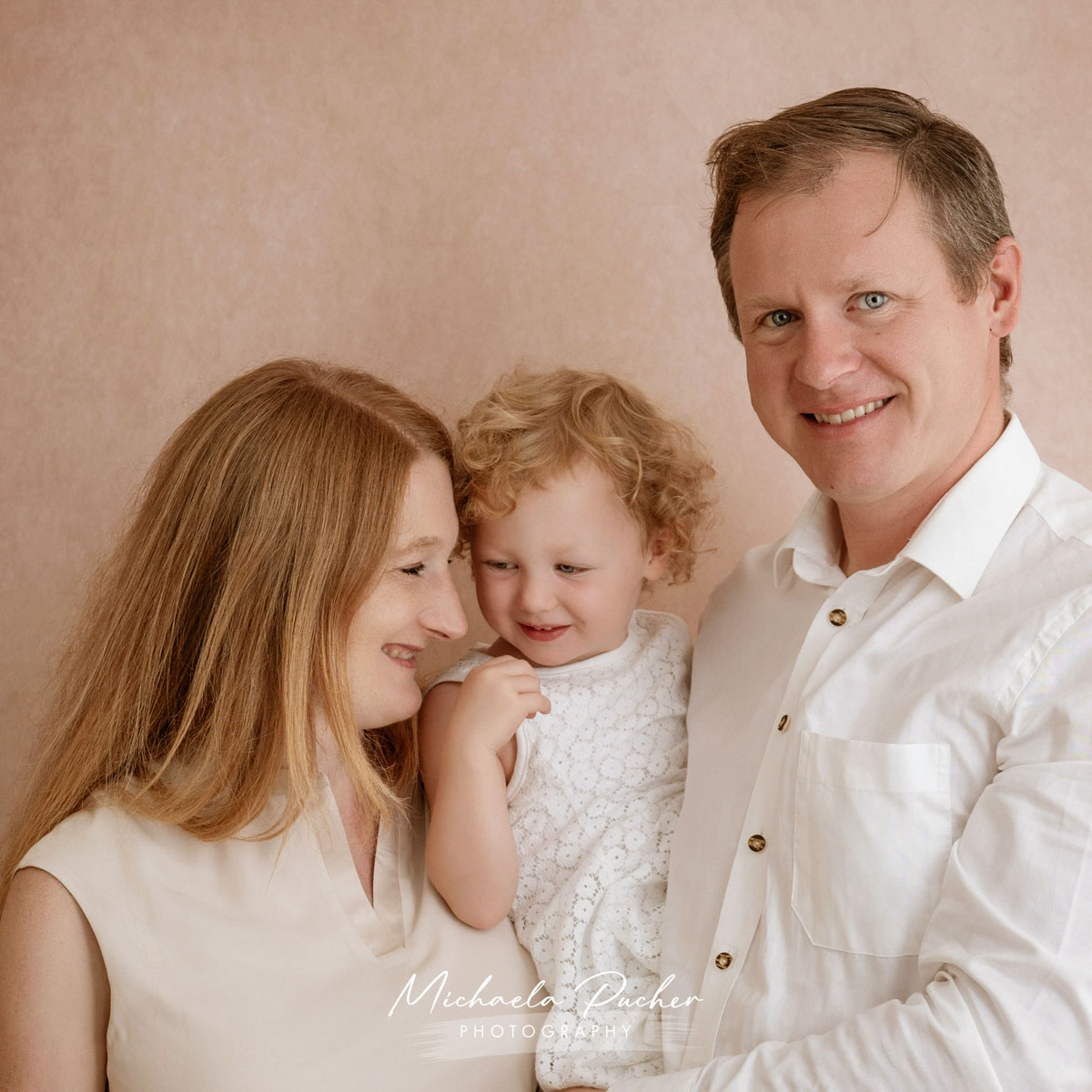 Eltern halten ihr kleines Kind im Arm; alle drei lächeln sich liebevoll an in einer hellen, sanften Studioumgebung.